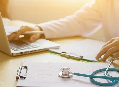 a doctor working on a laptop while looking at papers on a clipboard and a stethoscope in the foreground