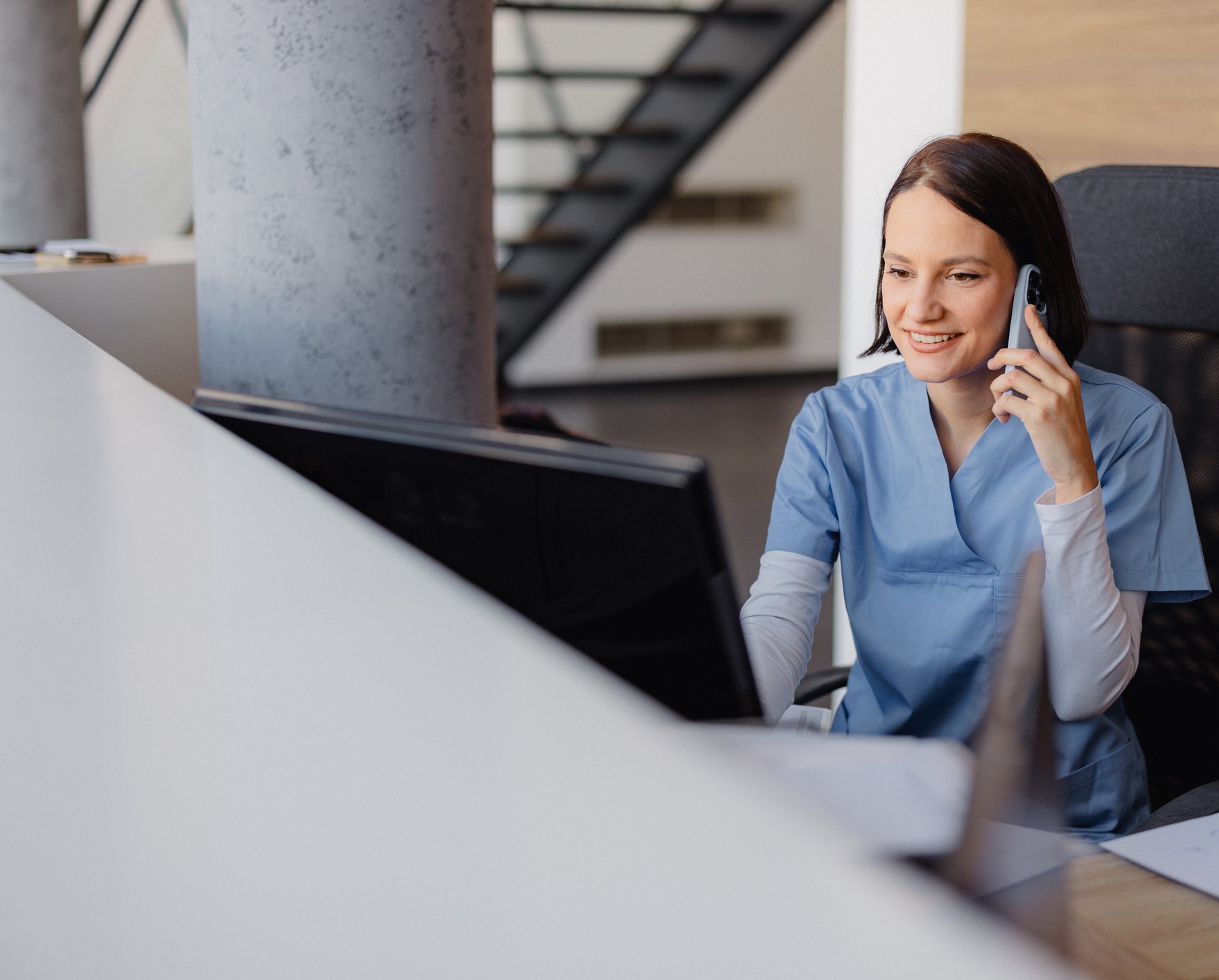 female health provider wearing blue scrubs talking on the phone while sitting in front of a computer