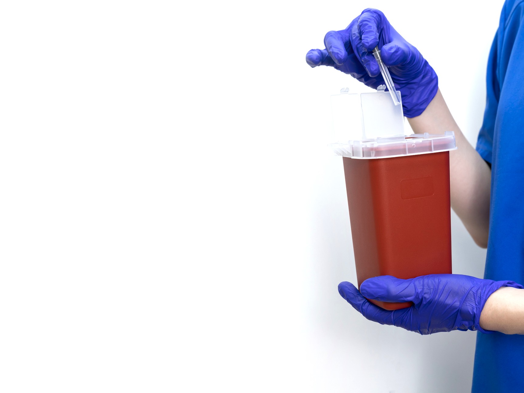 provider wearing gloves putting a syringe into a red medical waste collection container in an all white background
